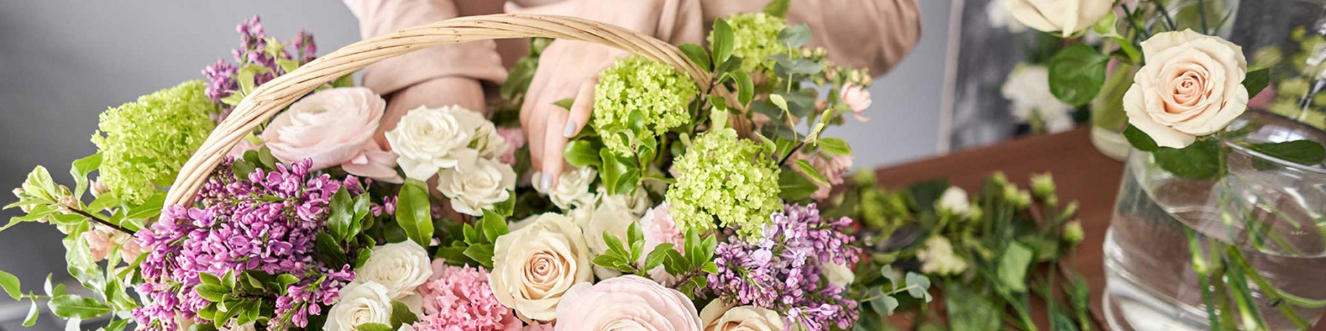 En person holder en kurv fyldt med farverige blomster, herunder roser, ranunkler og liljekonvaller. Der er også flere friske blomster på bordet, og stemningen er lys og indbydende.